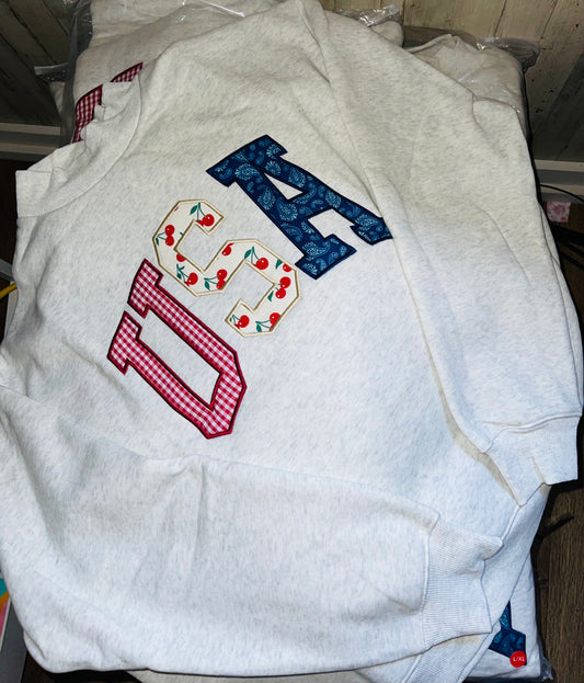 Light gray crewneck sweatshirt laid flat on Featuring large “USA” letters in red, white, and blue patterned fabric across the front.