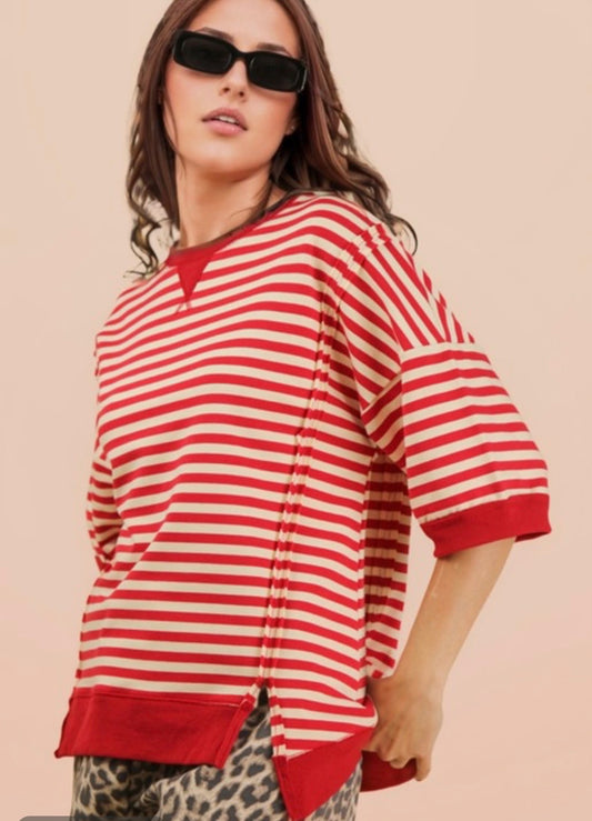 A side-angle view of a woman wearing a red and cream striped oversized top and dark rectangular sunglasses. The image highlights the shirt's dropped shoulder seam and a vertical side slit at the hem. She has one hand tucked into the pocket of her leopard print pants. The lighting is soft against a neutral beige background.
