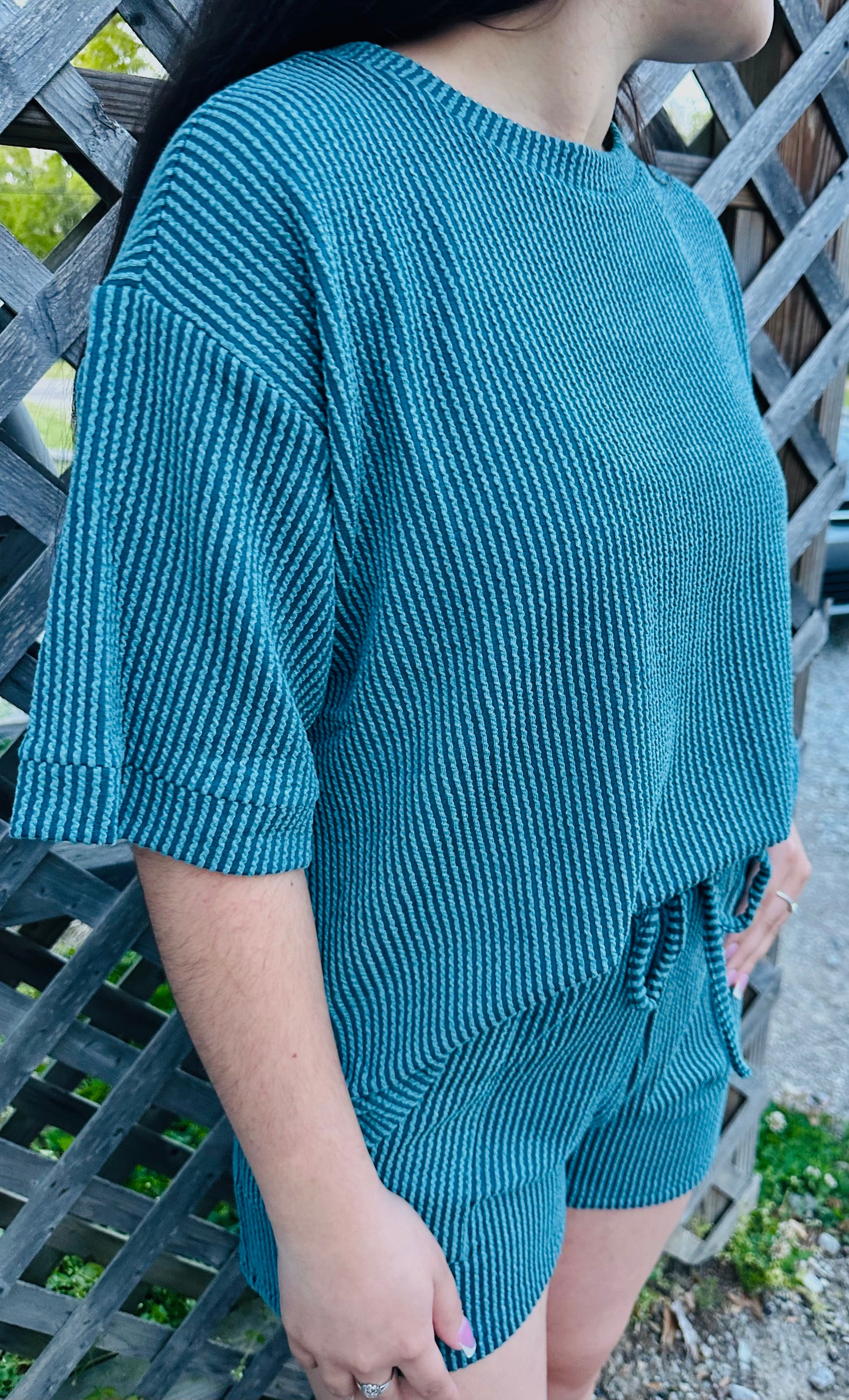 A person modeling a two-piece lounge set made of textured knit fabric. The set includes a loose short-sleeve top and matching drawstring shorts. It is shown in a dark Teal color. The model is standing outdoors against a wooden lattice, displaying the relaxed fit and soft texture of the outfit.