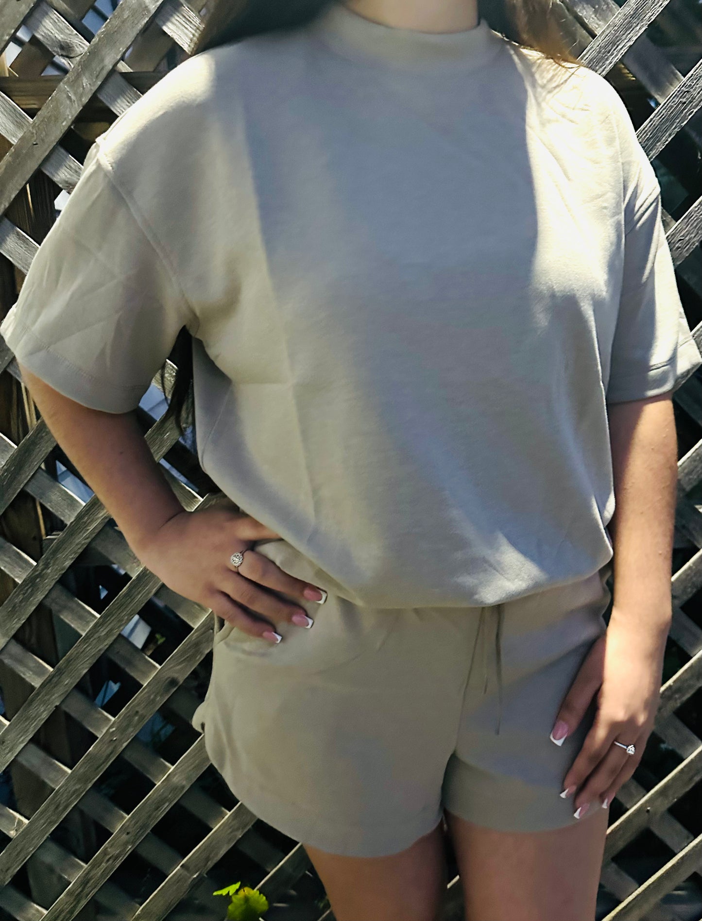 Model wearing a light beige two-piece lounge set with a loose short-sleeve top and matching drawstring shorts, standing outdoors in front of a wooden lattice.