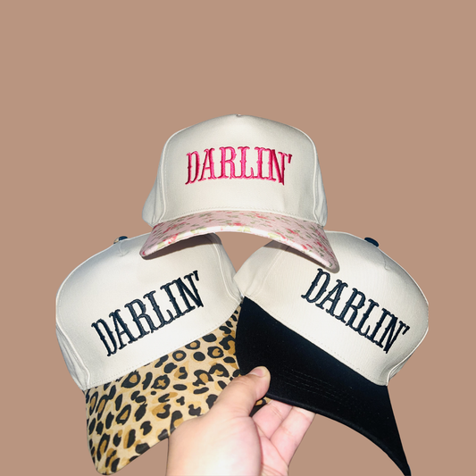 Three white caps with 'Darlin' text on a beige background. One had floral with pink Darlin' letters. Another has Leopard Print with Black letters that say Darlin. The other is Black and white with Black letters that say "Darlin'" as well.