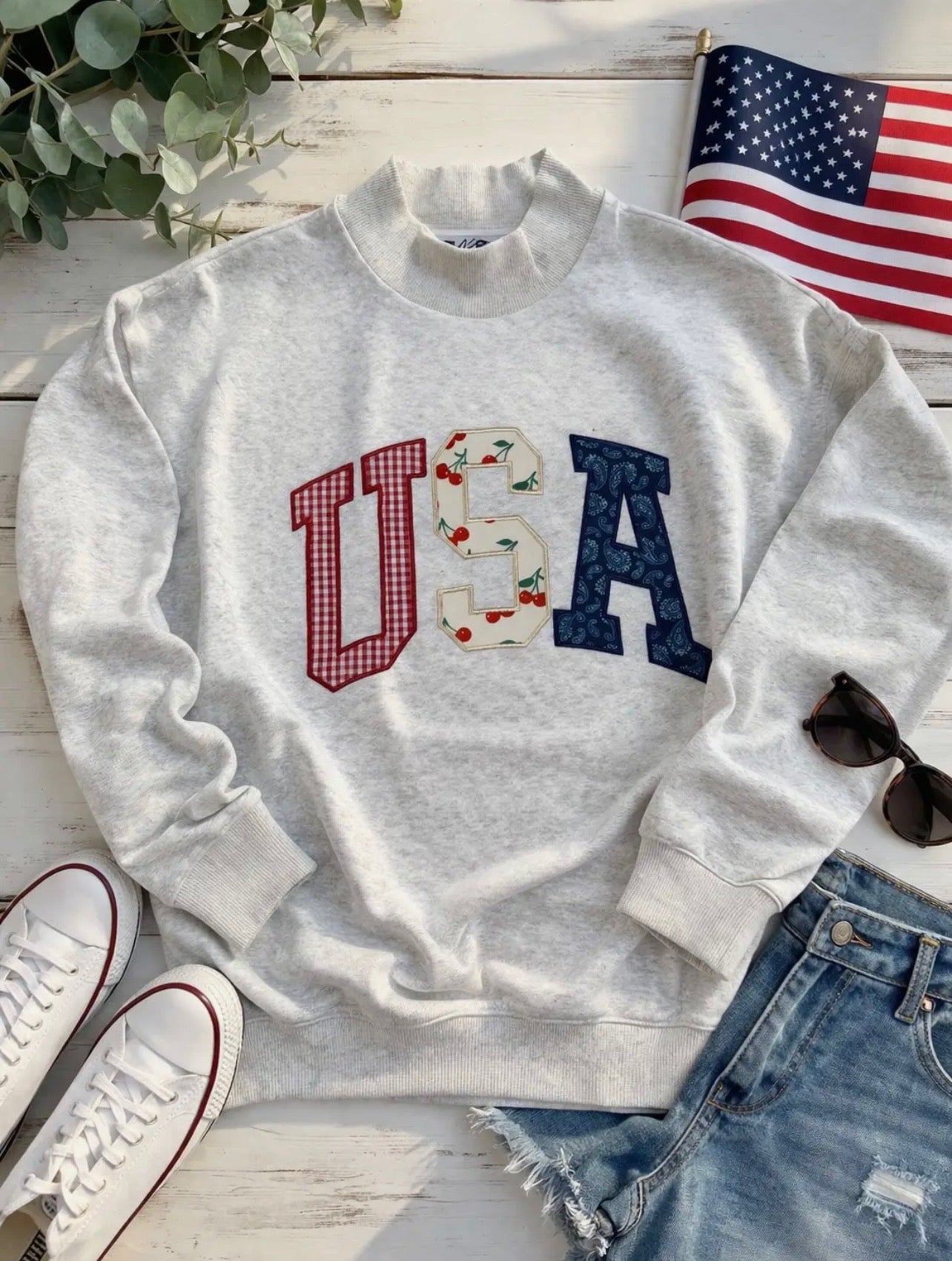 Light gray crewneck sweatshirt laid flat on a wooden surface, featuring large “USA” letters in red, white, and blue patterned fabric across the front. Styled with denim shorts, white sneakers, sunglasses, greenery, and a small American flag in the background