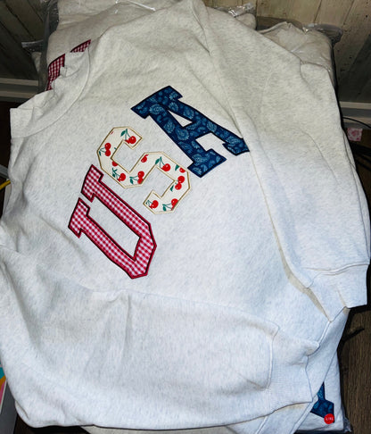 Light gray crewneck sweatshirt laid flat on Featuring large “USA” letters in red, white, and blue patterned fabric across the front. 