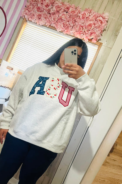 Women wearing a Light gray crewneck sweatshirt laid flat on Featuring large “USA” letters in red, white, and blue patterned fabric across the front. 