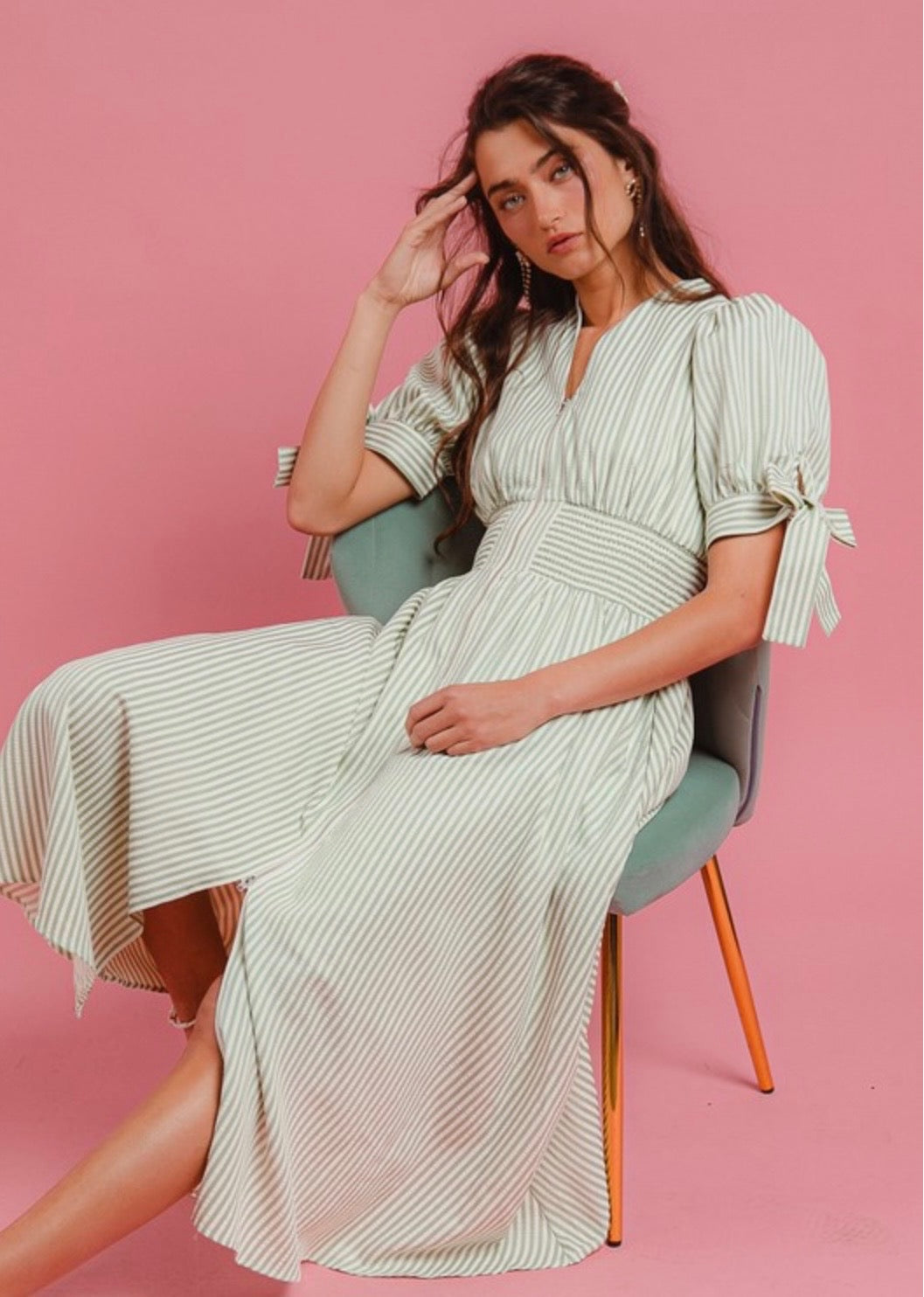 Woman wearing a sage green and white striped midi dress with puff sleeves and tie details, seated on a chair against a pink background.