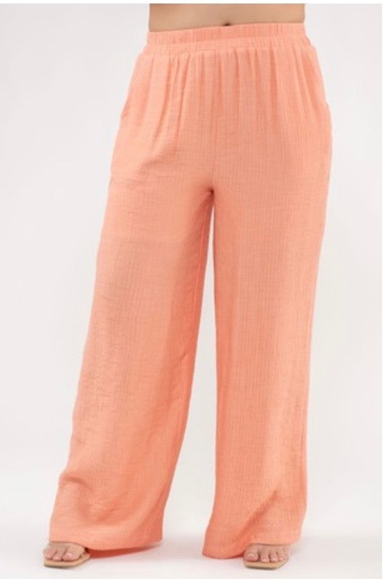 A studio shot showing the lower half of a model standing against a plain white background, wearing bright peach-colored, wide-leg pants. The pants are made of a lightweight, crinkled fabric and feature a gathered elastic waistband. The model is wearing minimalist square-toe sandals.