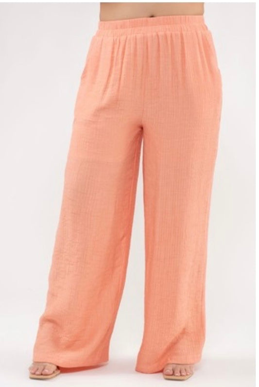 A studio shot showing the lower half of a model standing against a plain white background, wearing bright peach-colored, wide-leg pants. The pants are made of a lightweight, crinkled fabric and feature a gathered elastic waistband. The model is wearing minimalist square-toe sandals.