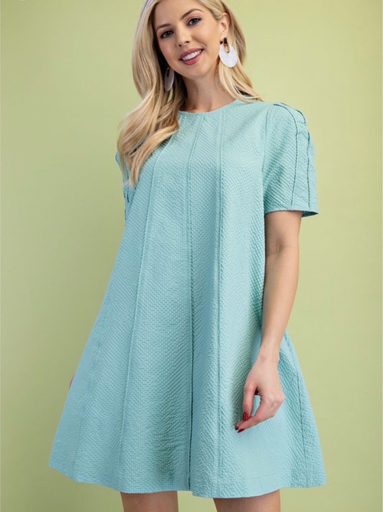 A person modeling a light mint green, short-sleeved dress. The dress is made of a highly textured, puckered fabric and features a high round neckline, slight gathering at the shoulders, and two vertical seams running down the front. Person has blonde hair and is wearing white heels and is standing in a green background.