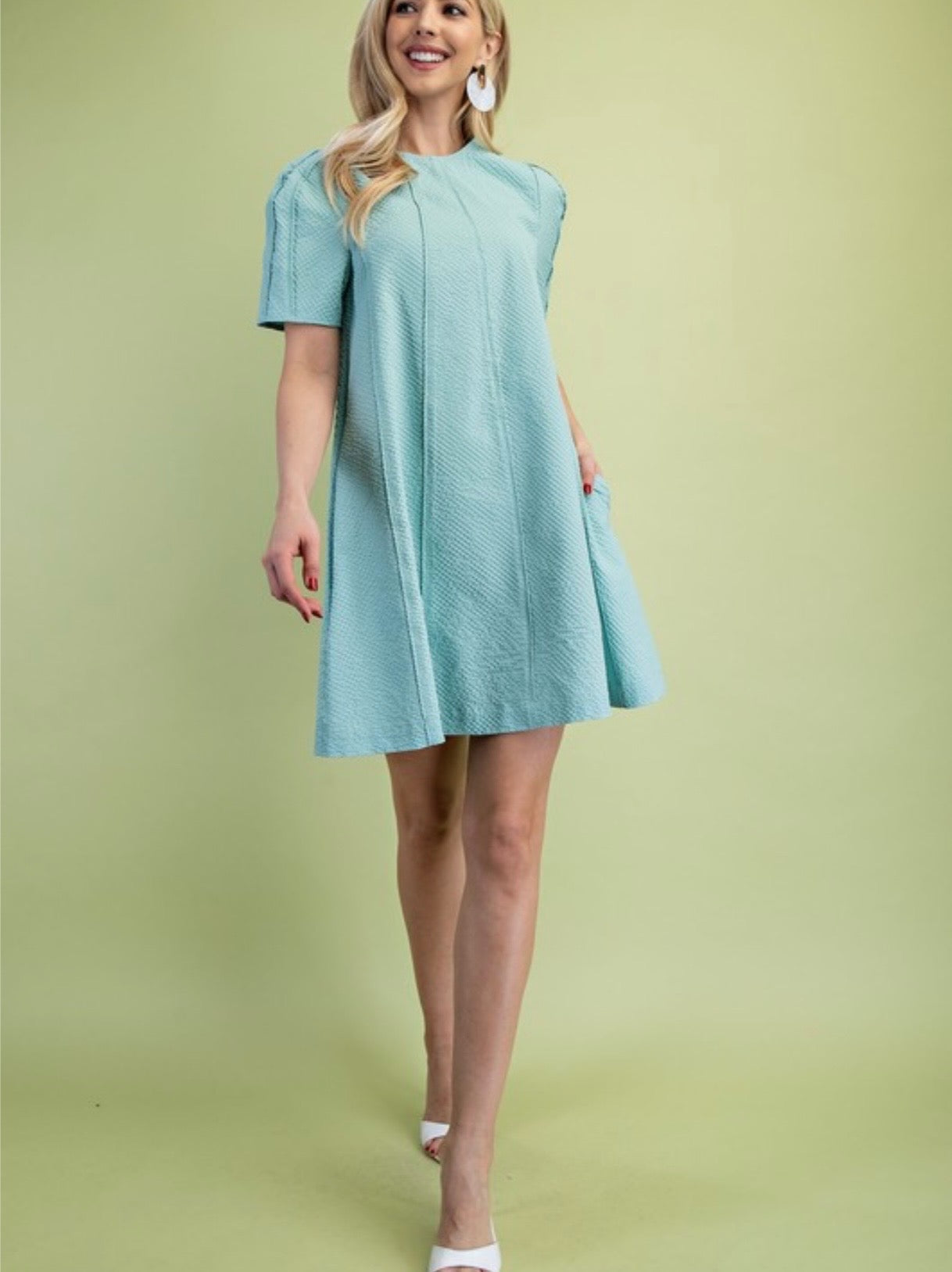 A person modeling a light mint green, short-sleeved dress. The dress is made of a highly textured, puckered fabric and features a high round neckline, slight gathering at the shoulders, and two vertical seams running down the front. Person has blonde hair and is wearing white heels and is standing in a green background.