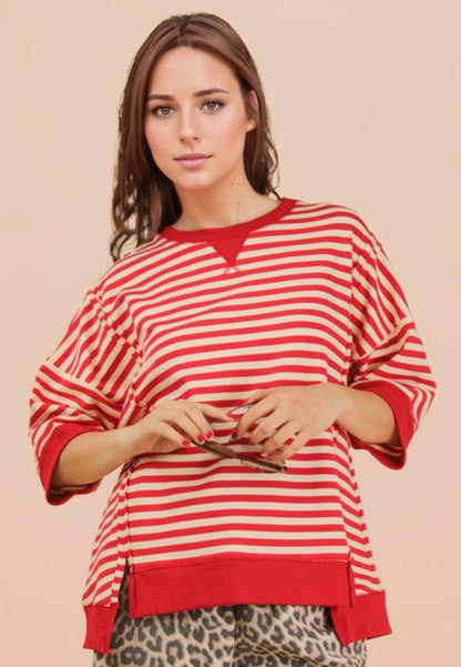 A medium shot of a woman with long brown hair wearing an oversized, red and cream horizontally striped short-sleeved sweatshirt. The top features a solid red crew neck with a V-stitch detail and a thick red banded hem. She is holding brown sunglasses in front of her. Below the shirt, leopard print bottoms are partially visible against a solid tan background.