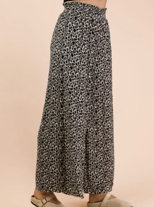 A side-profile shot from the waist down of a person wearing high-waisted, wide-leg trousers. The pants feature a dense, small-scale leopard print in grey and black. The waistband is wide and gathered with a ruffled top edge. The person is wearing cream-colored mesh slide sandals with a buckle strap. The background is a solid, neutral tan.