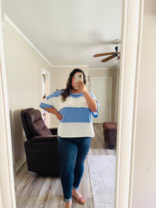 Person wearing a white and blue color block striped oversized short sleeve top with a relaxed fit, paired with dark denim jeans AND taking a mirror selfie in a room with a couch and ceiling fan.