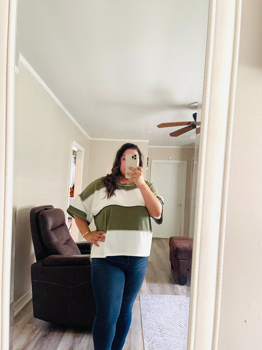 Person wearing a white olive color block striped oversized short sleeve top with a relaxed fit, paired with dark denim jeans AND taking a mirror selfie in a room with a couch and ceiling fan.