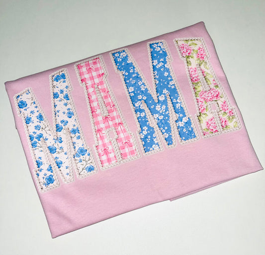 Pink fabric with embroidered letters 'N' and 'M' using floral and checkered patterns on a white background.