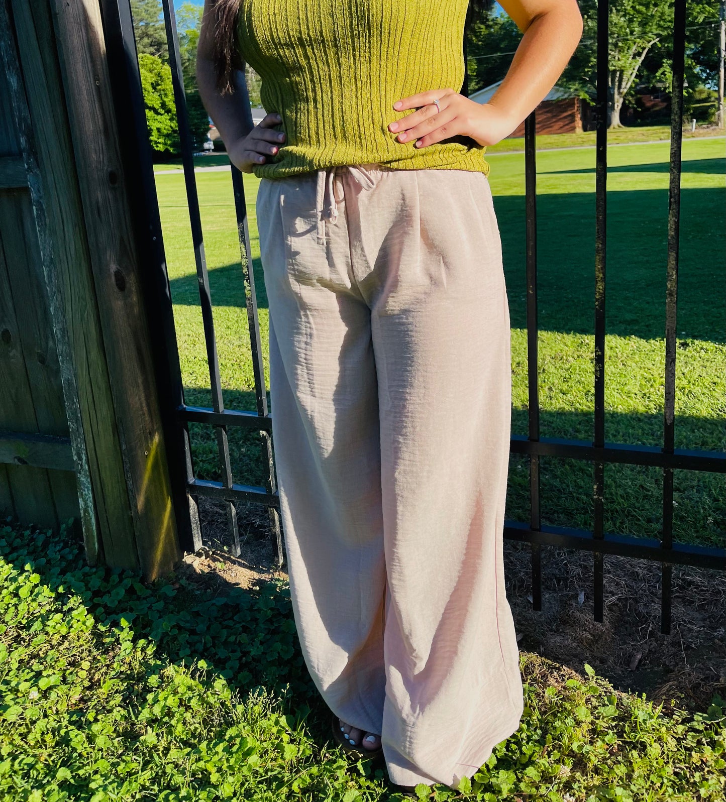 A person outdoors modeling light beige, flowy wide-leg pants. The pants are made of a textured, crinkled fabric and feature an elastic waistband with a front drawstring tie and subtle pleating. The person has long dark hair and is also wearing a ribbed, olive-green sleeveless top.