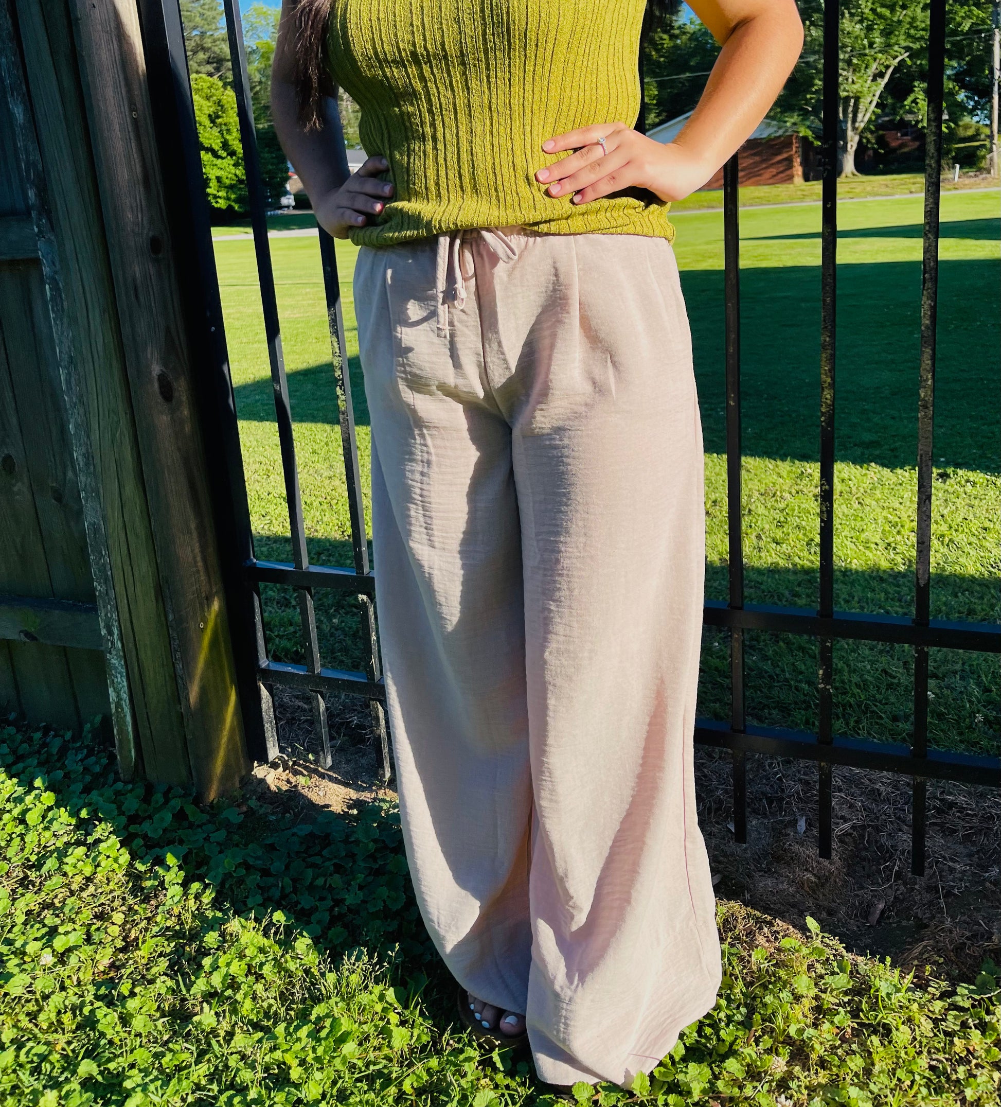 A person outdoors modeling light beige, flowy wide-leg pants. The pants are made of a textured, crinkled fabric and feature an elastic waistband with a front drawstring tie and subtle pleating. The person has long dark hair and is also wearing a ribbed, olive-green sleeveless top.