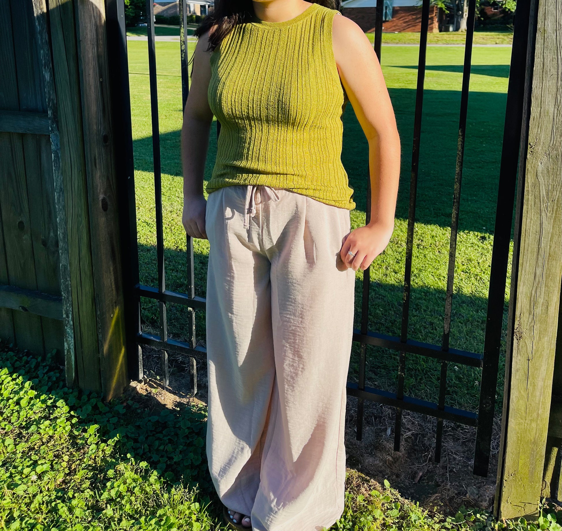 A person standing outdoors in front of a black metal fence on a sunny day. They are wearing a fitted, olive green ribbed knit sleeveless top and light beige, wide-leg crinkle pants. Their hands are resting casually near the waistband of the pants.
