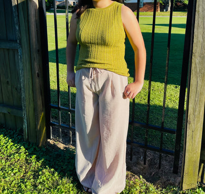 A person standing outdoors in front of a black metal fence on a sunny day. They are wearing a fitted, olive green ribbed knit sleeveless top and light beige, wide-leg crinkle pants. Their hands are resting casually near the waistband of the pants.
