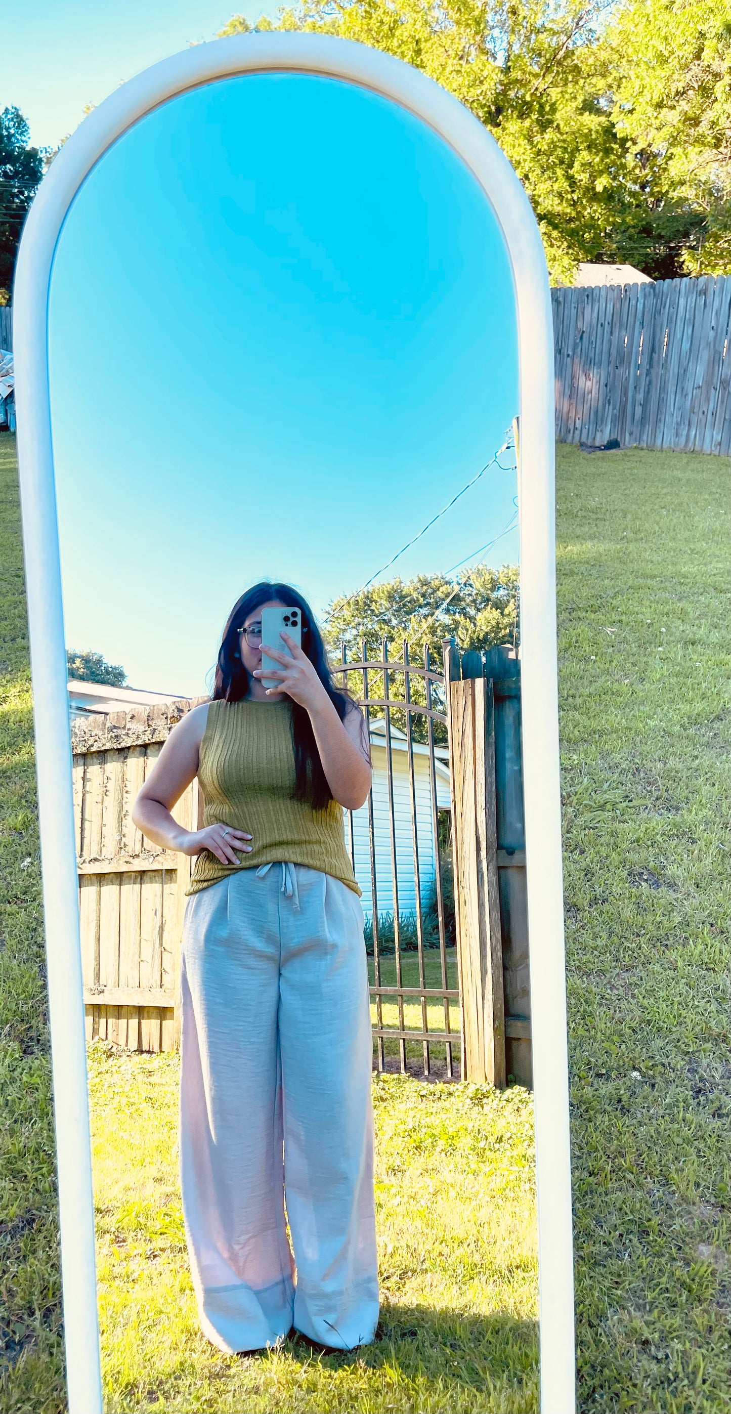 A full-length outdoor mirror selfie of a person modeling light beige, crinkled wide-leg pants with a drawstring waist and a ribbed olive-green sleeveless top. The person's reflection is captured in a tall, white arched mirror standing on a grassy lawn, with a clear blue sky, trees, and a fence visible in the background reflection.