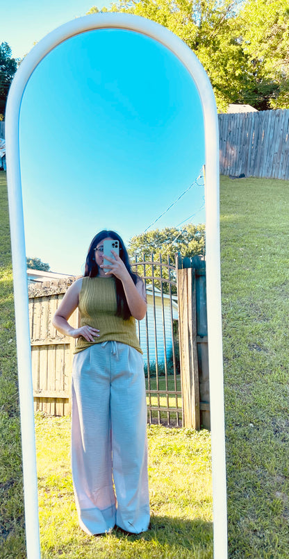 A full-length outdoor mirror selfie of a person modeling light beige, crinkled wide-leg pants with a drawstring waist and a ribbed olive-green sleeveless top. The person's reflection is captured in a tall, white arched mirror standing on a grassy lawn, with a clear blue sky, trees, and a fence visible in the background reflection.
