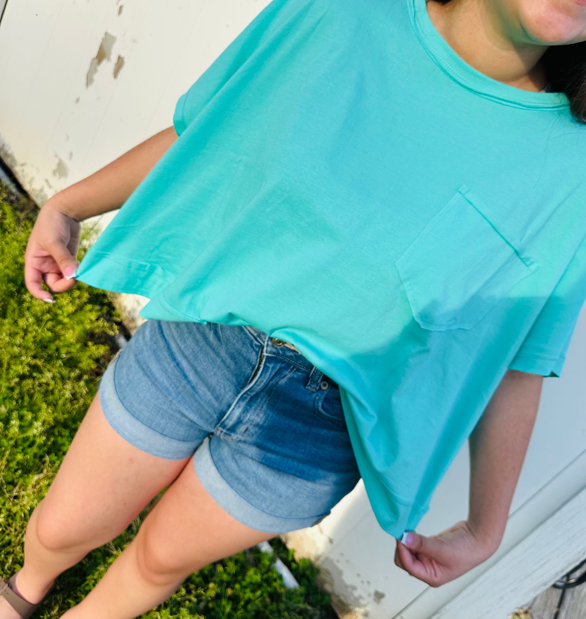 A person modeling a short-sleeve, loose-fit t-shirt with a small chest pocket. The shirt has a relaxed, slightly cropped silhouette and is shown in bright coral and aqua colors. It is paired with rolled denim shorts. The model is standing outdoors in natural light, showing both front and back views of the shirt, highlighting its soft drape and casual style.