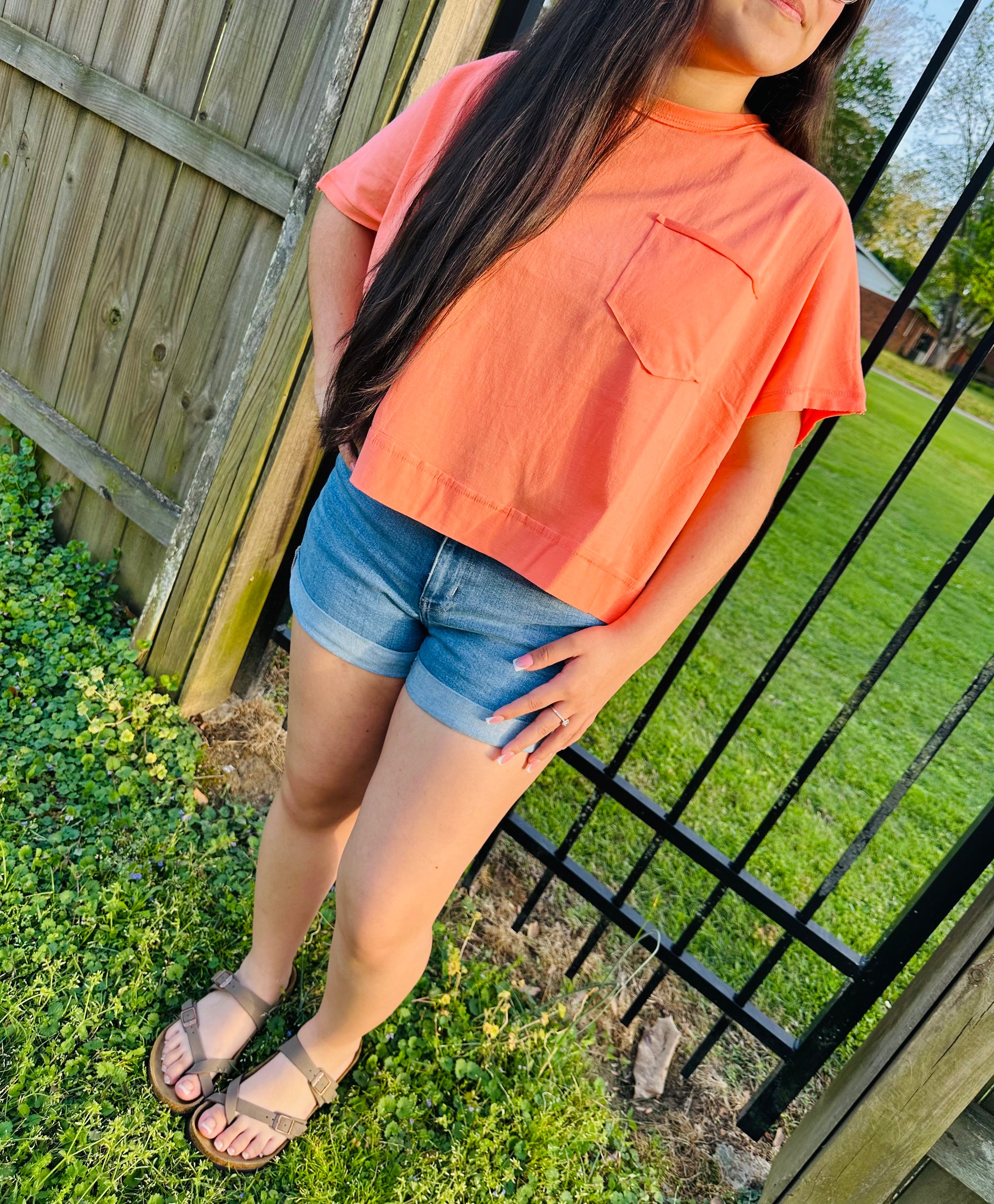 Person wearing an orange shirt and denim shorts standing outdoors near a fence and grass.