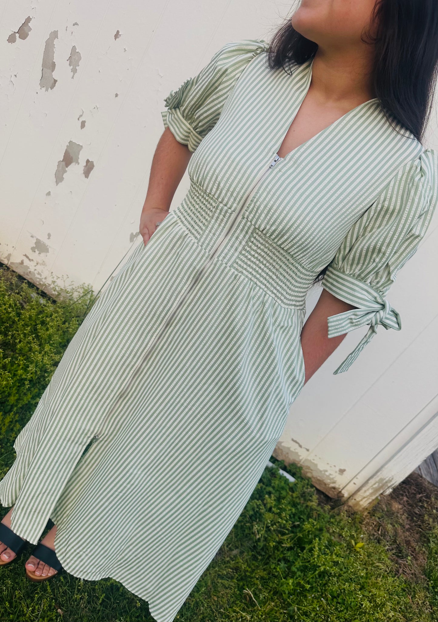 Close-up of a person wearing a sage green striped midi dress with a front zipper, smocked waist, and short puff sleeves with tie accents.