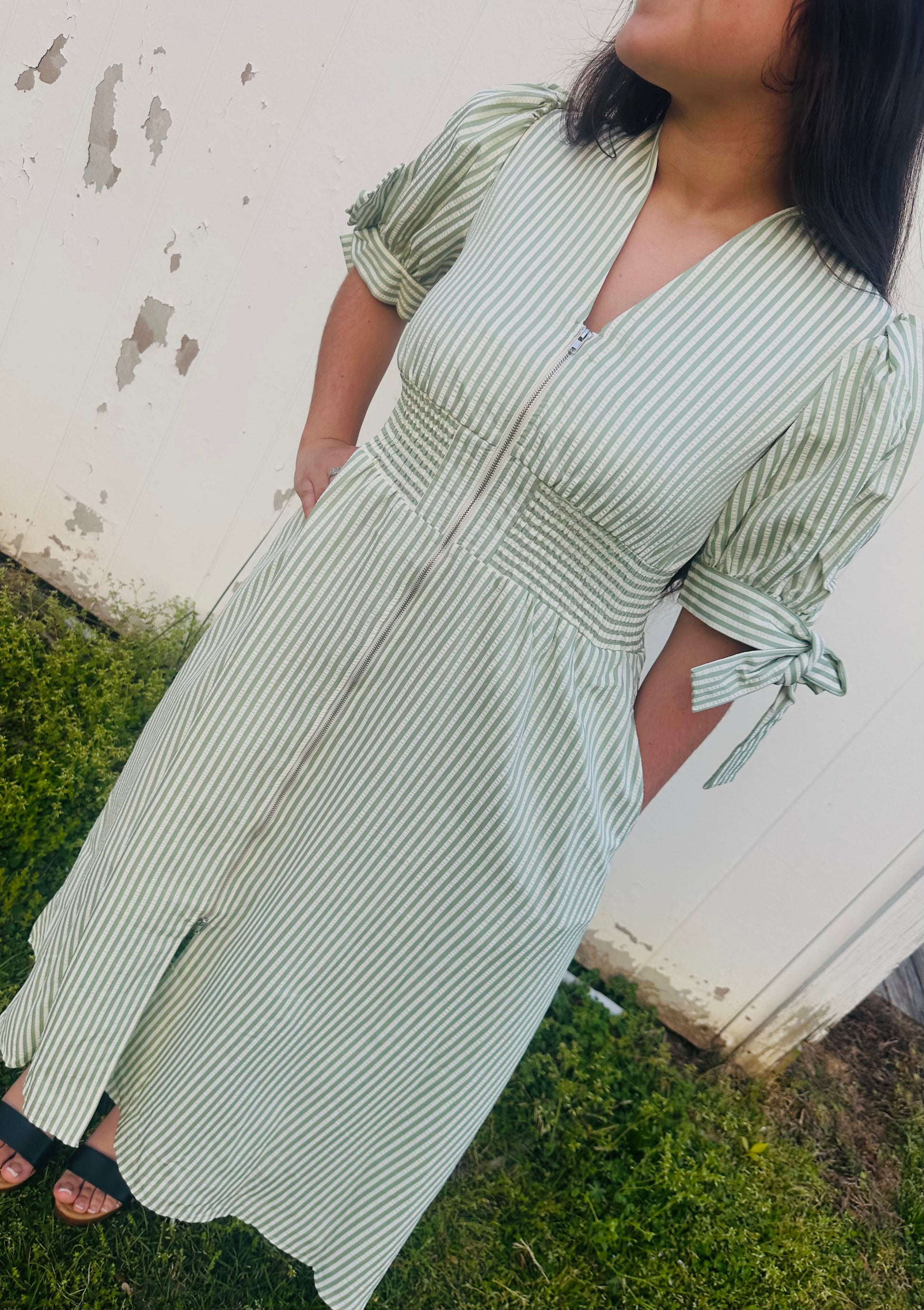 Close-up of a person wearing a sage green striped midi dress with a front zipper, smocked waist, and short puff sleeves with tie accents.