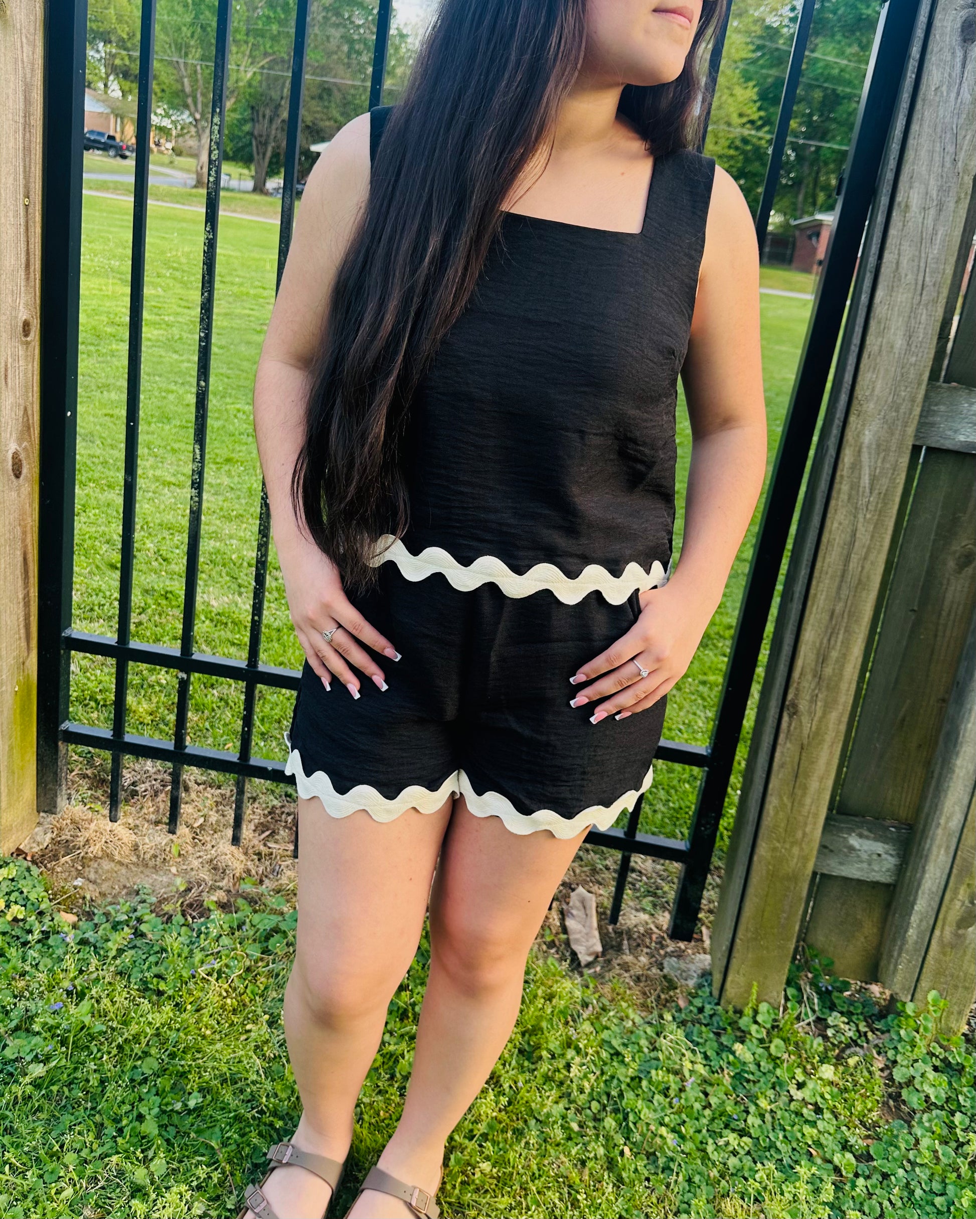 Model wearing a black two-piece lounge set with a sleeveless top and matching shorts featuring light-colored wavy trim, standing outdoors near a wooden fence with hands in her pocket.