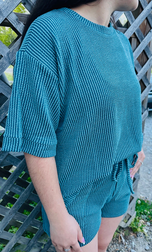 A person modeling a two-piece lounge set made of textured knit fabric. The set includes a loose short-sleeve top and matching drawstring shorts. It is shown in a dark Teal color. The model is standing outdoors against a wooden lattice, displaying the relaxed fit and soft texture of the outfit.