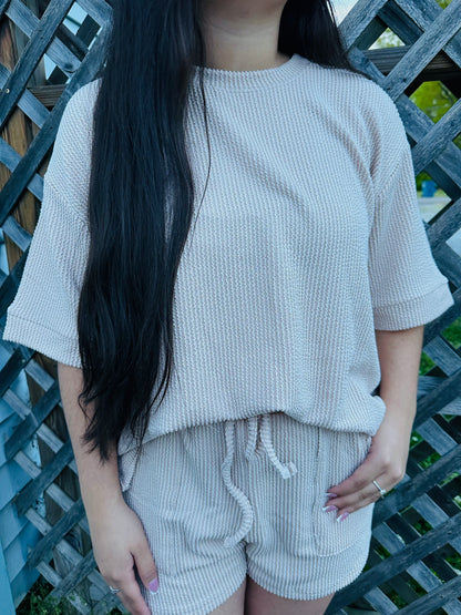 A person modeling a two-piece lounge set made of textured knit fabric. The set includes a loose short-sleeve top and matching drawstring shorts. It is shown in neutral cream The model is standing outdoors against a wooden lattice, displaying the relaxed fit and soft texture of the outfit.