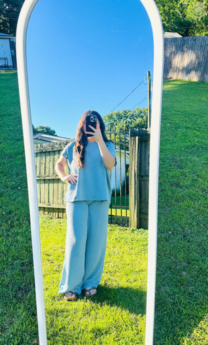 Model wearing a light blue ribbed lounge set featuring a loose short-sleeve top and wide-leg pants, photographed outdoors in a full-length mirror on grass.
