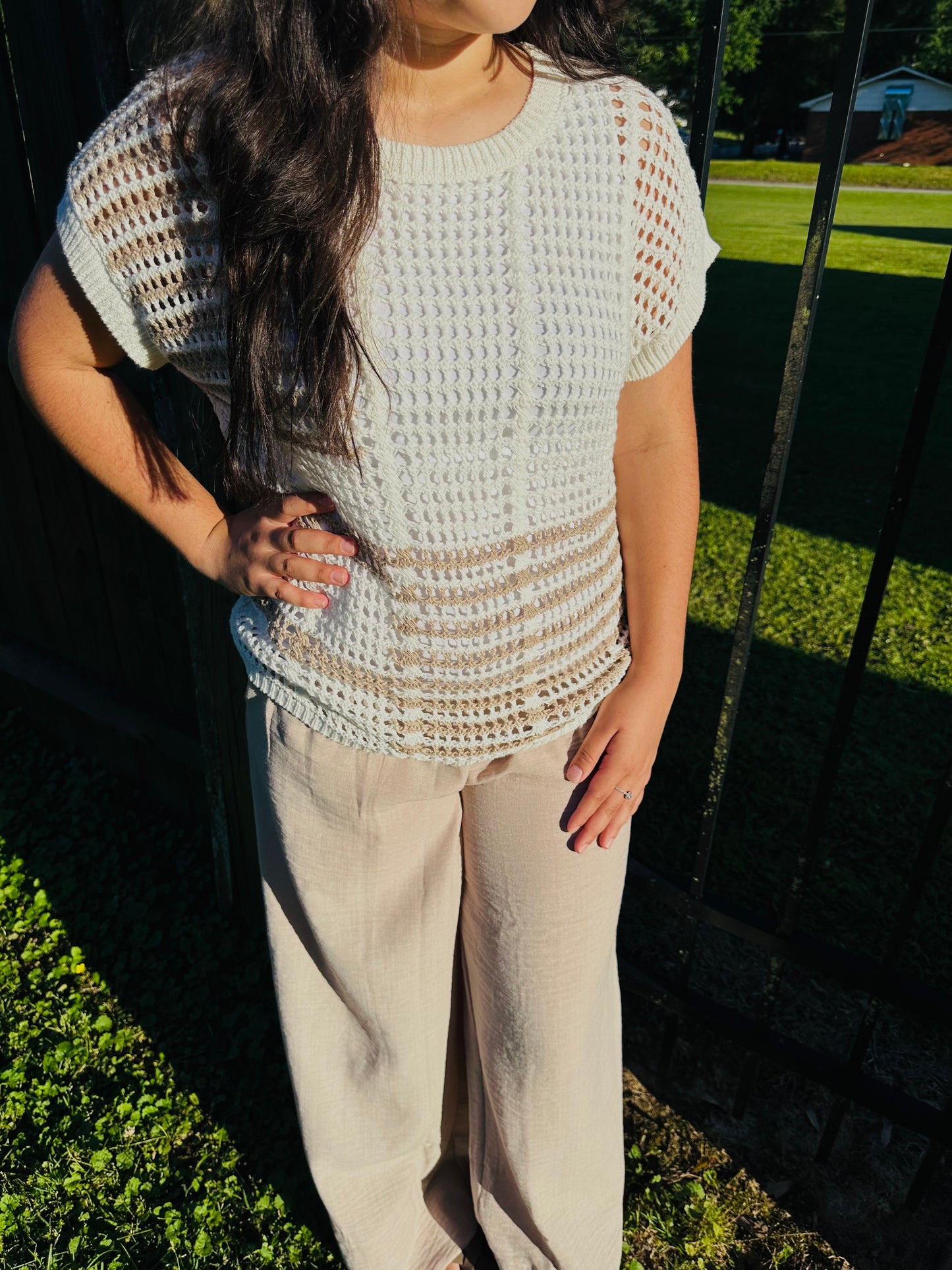 A person standing outdoors next to a black metal fence, modeling a white, open-knit short-sleeved top. The top features a slightly sheer crochet-like pattern and horizontal tan stripes across the lower section. The top is layered over a white camisole and paired with light beige wide-leg pants. The person has their right hand resting on their hip.