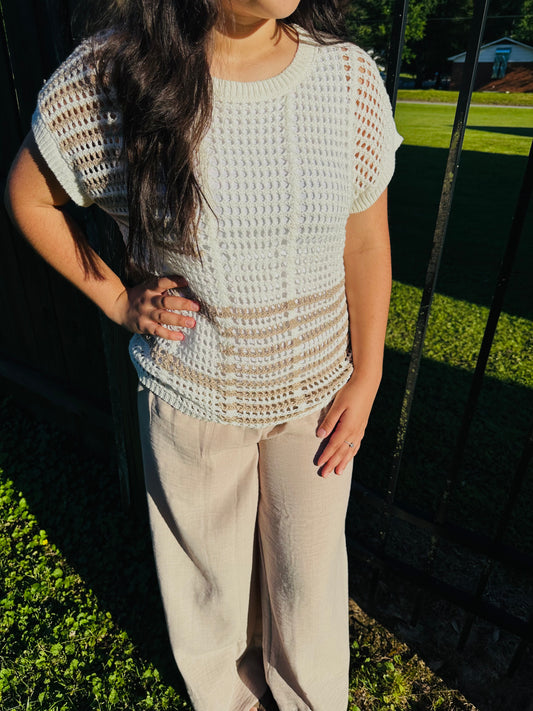 A person standing outdoors next to a black metal fence, modeling a white, open-knit short-sleeved top. The top features a slightly sheer crochet-like pattern and horizontal tan stripes across the lower section. The top is layered over a white camisole and paired with light beige wide-leg pants. The person has their right hand resting on their hip.