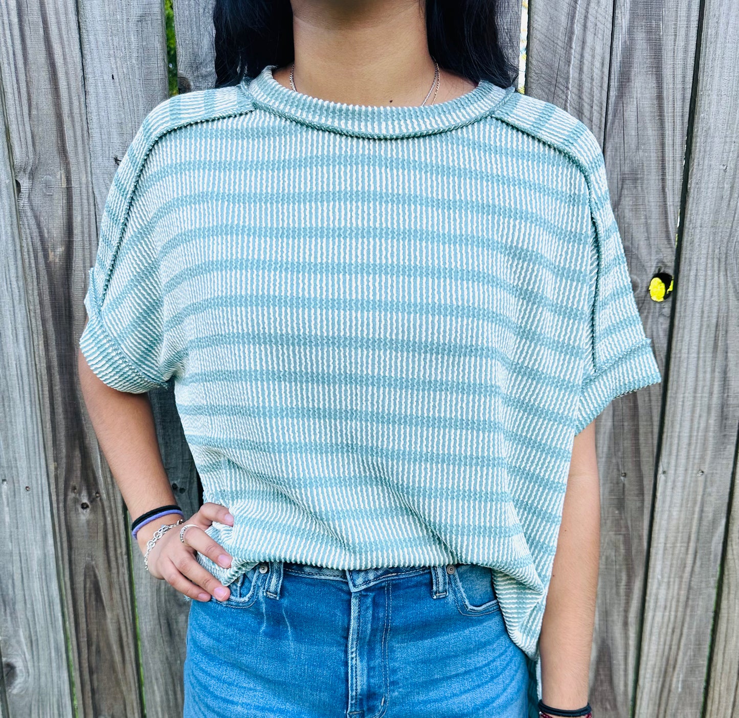 A person standing outdoors in front of a weathered wooden fence, modeling an oversized, short-sleeved top with horizontal teal and white stripes. The top features a highly textured, puckered fabric, a rolled crew neckline, and prominent exposed seams along the dropped shoulders. The person is wearing the top slightly front-tucked into distressed blue denim shorts.