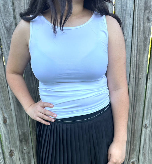 A person standing outdoors in front of a weathered wooden fence, modeling a fitted, solid white sleeveless tank top. The top features a high crew neckline and wide shoulder straps. The person is resting their hands on their hips and is also wearing a black pleated mini skirt.