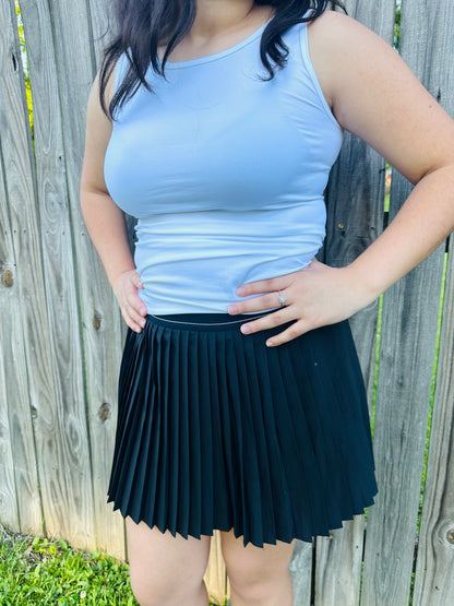 A person standing outdoors in front of a weathered wooden fence, modeling a fitted, solid white sleeveless tank top. The top features a high crew neckline and wide shoulder straps. The person is resting their hands on their hips and is also wearing a black pleated mini skirt.