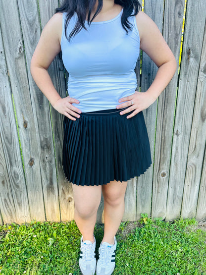 Pleated skirt paired with a white top on a model standing in a outdoor environment with grass and a fence in the background.