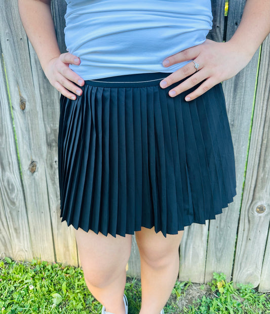 Pleated skirt paired with a white top on a model standing in a outdoor environment with grass and a fence in the background.