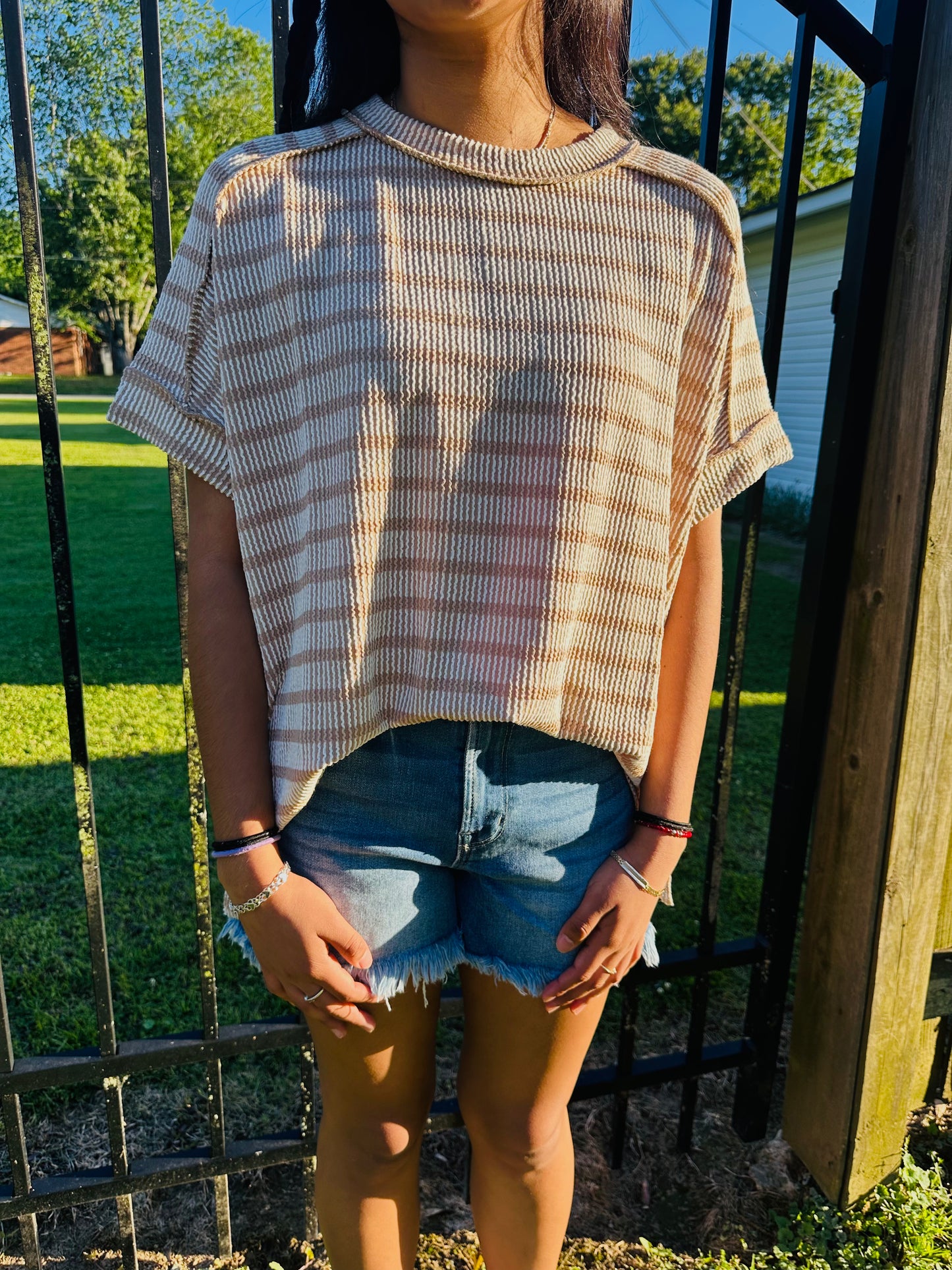 A person standing outdoors in front of a black metal fence, modeling a relaxed, oversized short-sleeved top with horizontal tan and white stripes. The top is made of a textured fabric with exposed shoulder seams and a rolled neckline. The person's arms are resting straight down at their sides, and they are wearing the top slightly front-tucked into cutoff denim shorts.