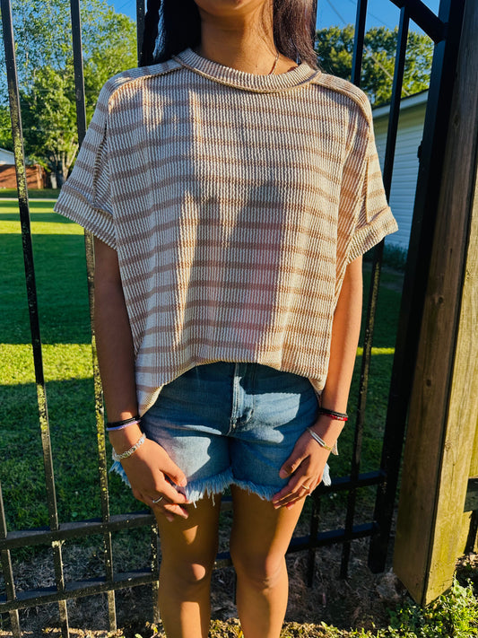 A person standing outdoors in front of a black metal fence, modeling a relaxed, oversized short-sleeved top with horizontal tan and white stripes. The top is made of a textured fabric with exposed shoulder seams and a rolled neckline. The person's arms are resting straight down at their sides, and they are wearing the top slightly front-tucked into cutoff denim shorts.