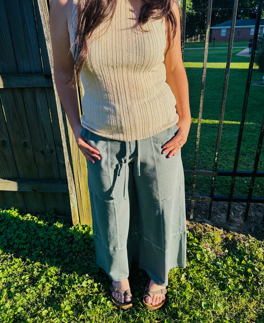 A person standing outdoors next to a black metal fence, modeling a fitted, light beige ribbed knit sleeveless top. The person has their hands resting on their pockets and is also wearing sage green, wide-leg pants with center-seam detailing