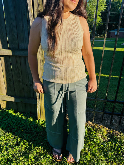 A person standing outdoors modeling relaxed-fit, sage green, wide-leg pants. The pants feature an elastic drawstring waist, large front patch pockets, and visible center-front seams running down each leg. The pants are paired with a textured beige sleeveless top.