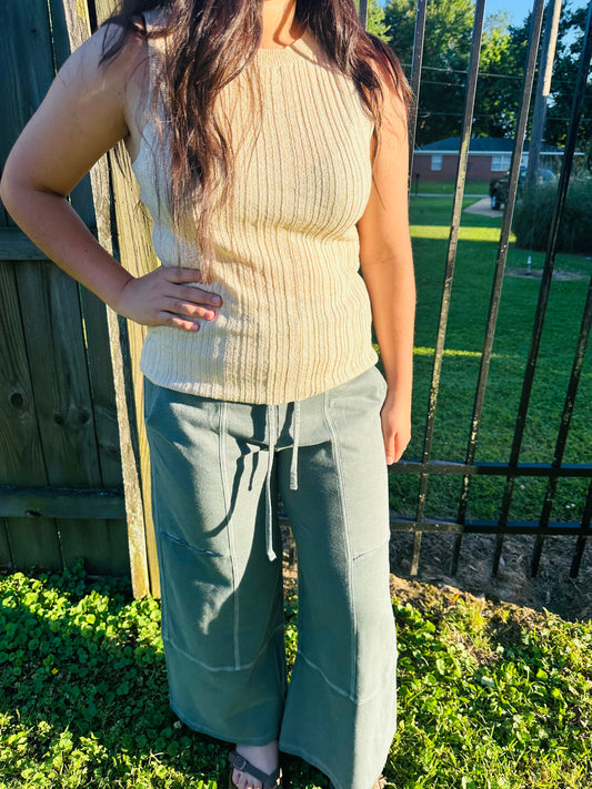 A person standing outdoors next to a black metal fence, modeling a fitted, light beige ribbed knit sleeveless top. The person has their right hand resting on their hip and is also wearing sage green, wide-leg pants with center-seam detailing.