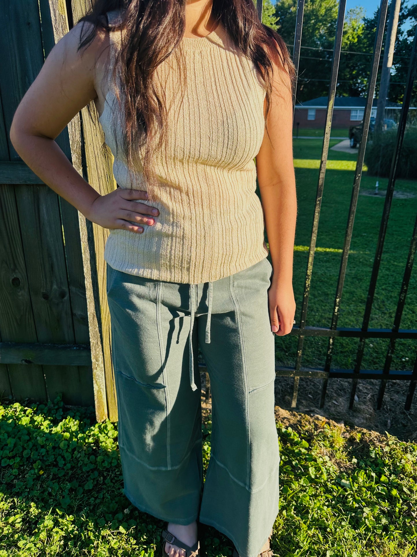 A person standing outdoors modeling relaxed-fit, sage green, wide-leg pants. The pants feature an elastic drawstring waist, large front patch pockets, and visible center-front seams running down each leg. The pants are paired with a textured beige sleeveless top.