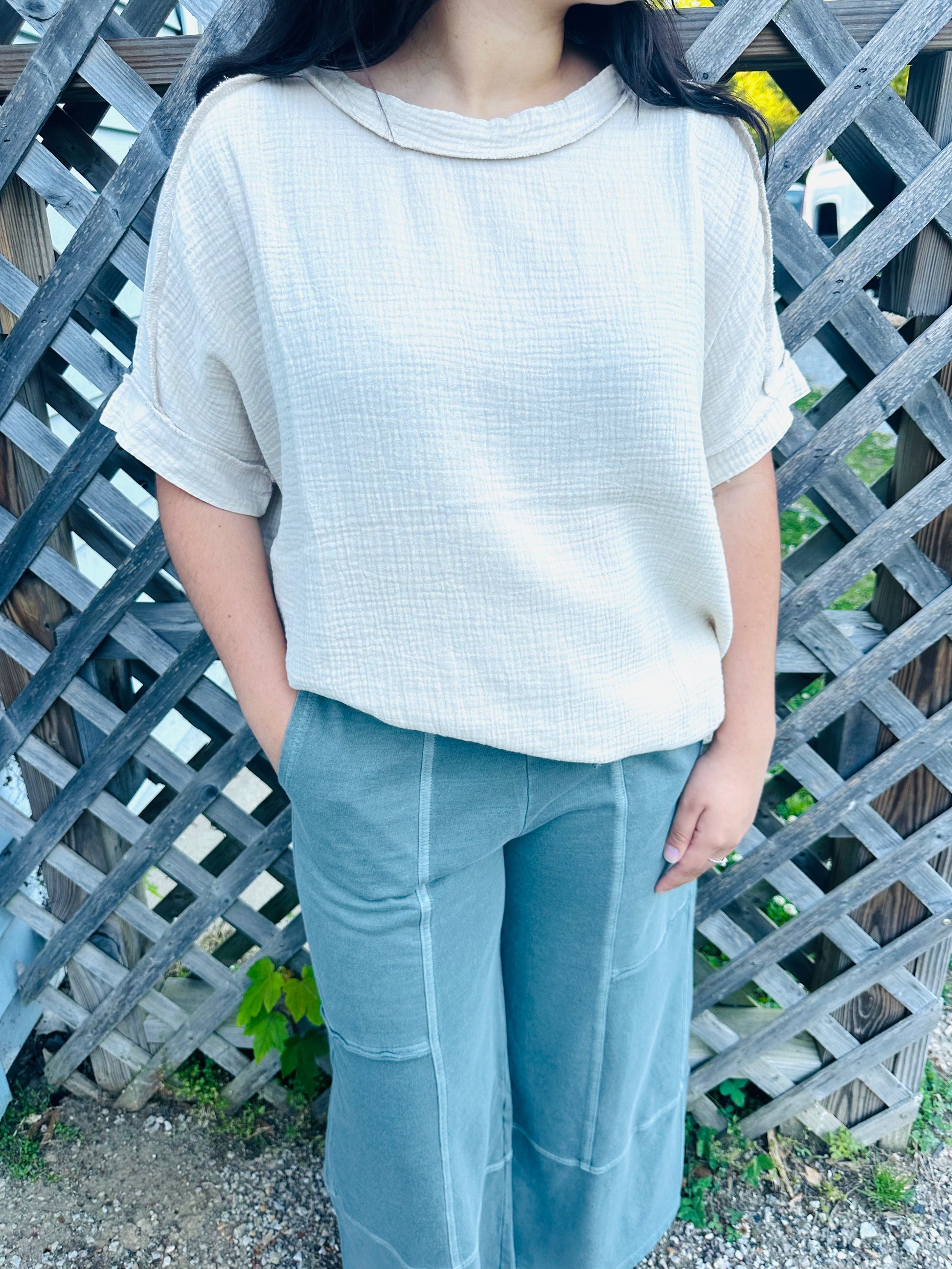 A person standing outdoors in front of a wooden lattice fence, modeling a relaxed, light beige short-sleeved top made of a textured, crinkled fabric. The top features a rolled crew neckline, drop shoulders, and rolled sleeve cuffs. The person has their hands resting in the front pockets of sage green wide-leg pants.