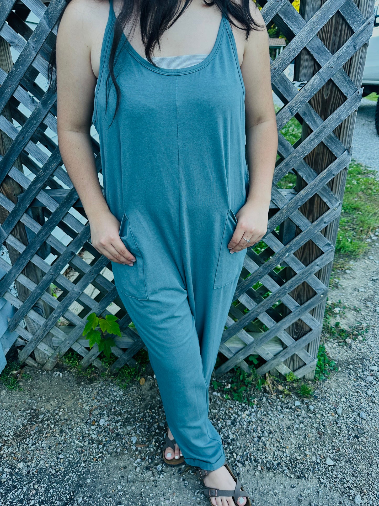 A person standing outdoors on a gravel path in front of a weathered wooden lattice fence, modeling a relaxed-fit, slate blue sleeveless jumpsuit. The person has their hands resting inside the jumpsuit's large front patch pockets. The jumpsuit features a scoop neckline, thin straps, and a center-front seam, and is layered over a light-colored top.