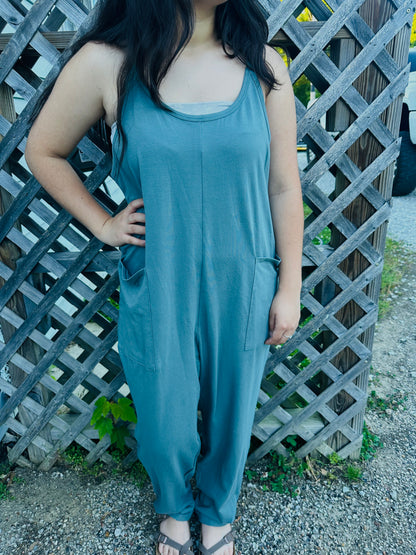 A person standing outdoors in front of a wooden lattice fence, modeling a slouchy, slate blue sleeveless jumpsuit. The jumpsuit has thin straps, a scoop neckline, large front patch pockets, and a center seam down the front. The person has their right hand resting casually on their hip.