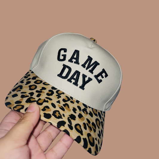 Cap with 'GAME DAY' text and leopard print brim held by a hand against a beige background